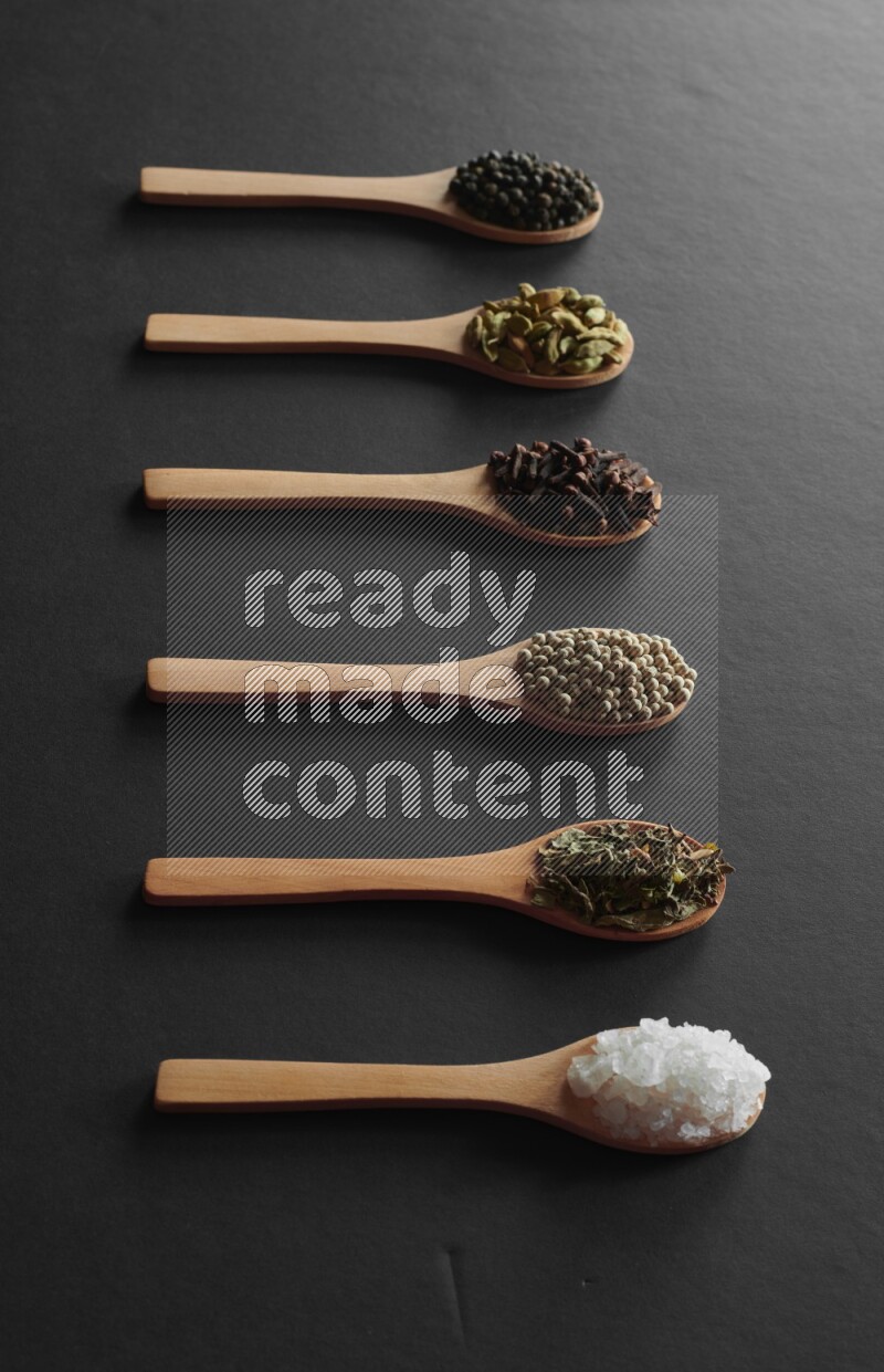wooden spoons filled with white peppers, cloves, cardamom, salt, black peppers and basil on black flooring