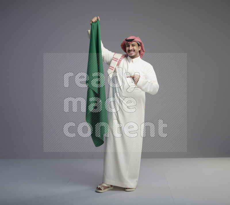 A saudi man standing wearing thob and red shomag holding big saudi flag on gray background