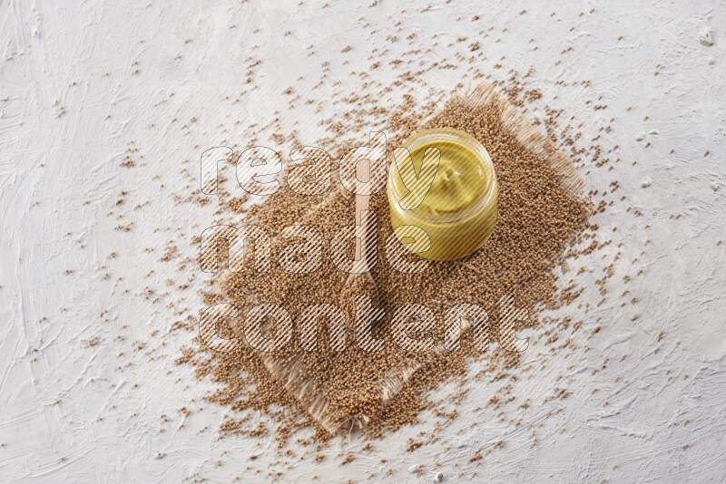 A glass jar full of mustard paste set on a burlap piece with a wooden spoon full of mustard seeds on a textured white flooring