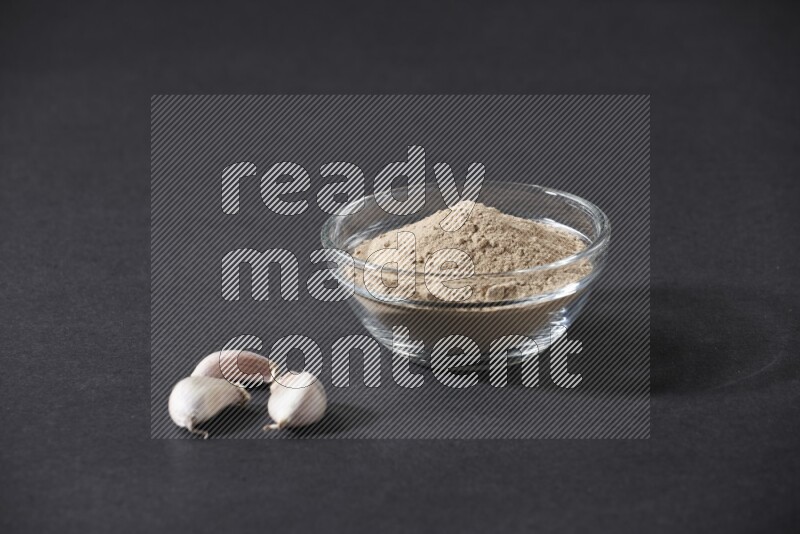 A glass bowl full of garlic powder with garlic bulb and some cloves beside it on a black flooring