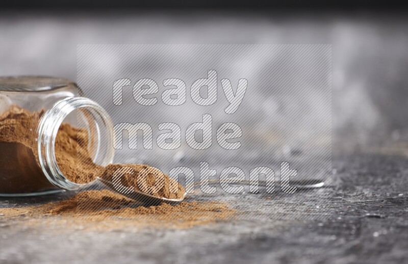 Herbal glass jar full of cinnamon powder flipped and a metal spoon on textured black background
