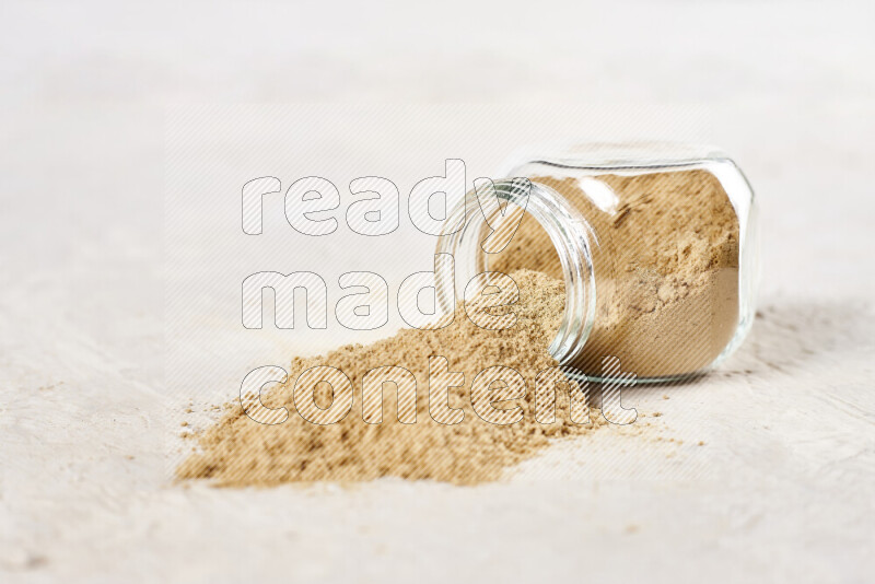 A glass jar full of ground ginger powder flipped with some spilling powder on white background