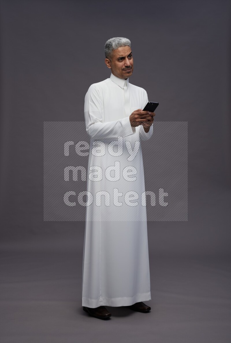 Saudi man wearing thob standing texting on phone on gray background