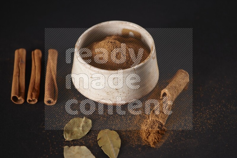 Cinnamon powder in a white pottery bowl and cinnamon sticks and laurel leaves on black background