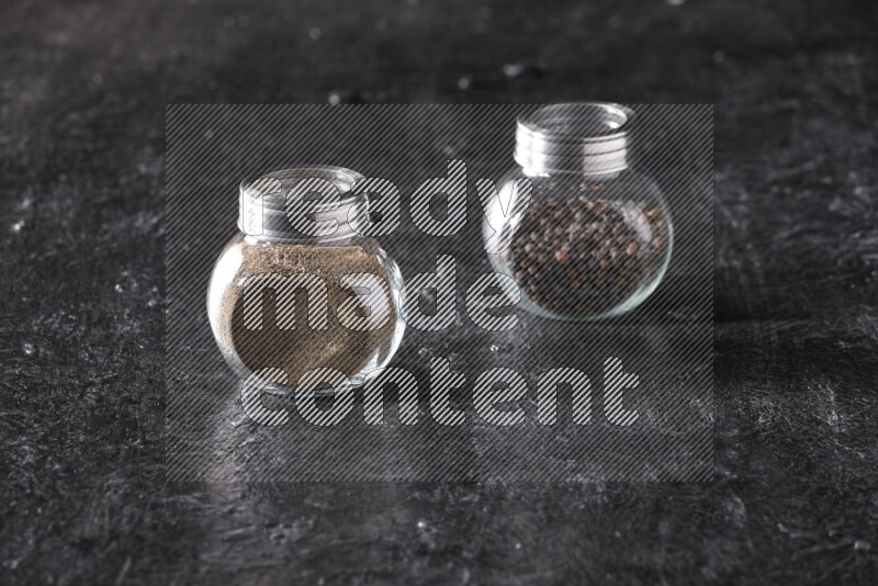 2 glass spice jars full of black pepper powder and black pepper beads on textured black flooring