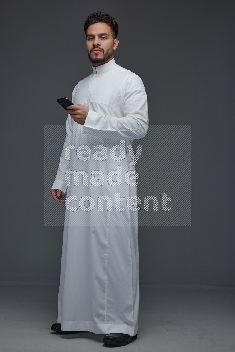 A Saudi man wearing Thobe standing and using his phone eye level on a gray background