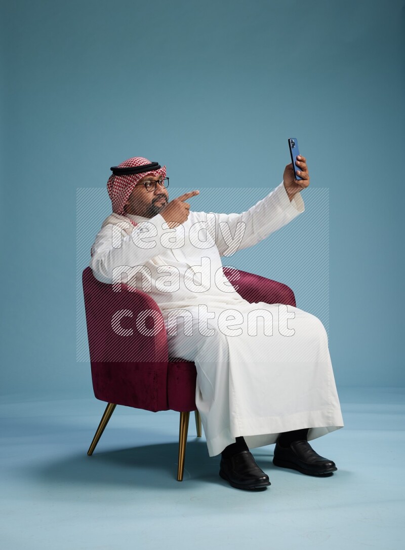 Saudi Man with shimag sitting on chair taking selfie on blue background