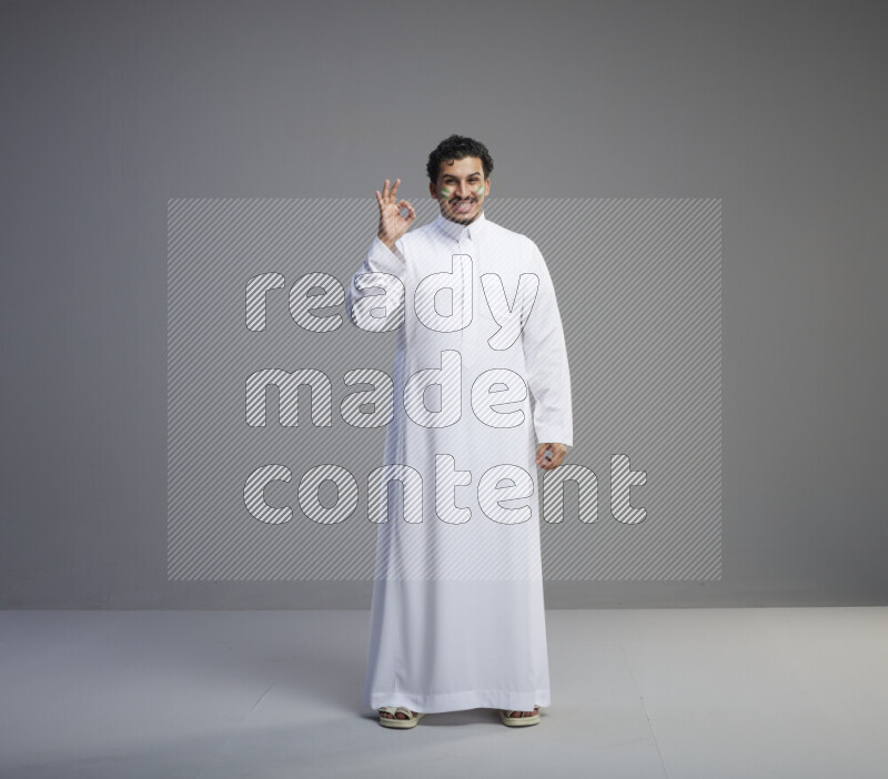 A Saudi man standing wearing thob with face painting interacting with the camera on gray background