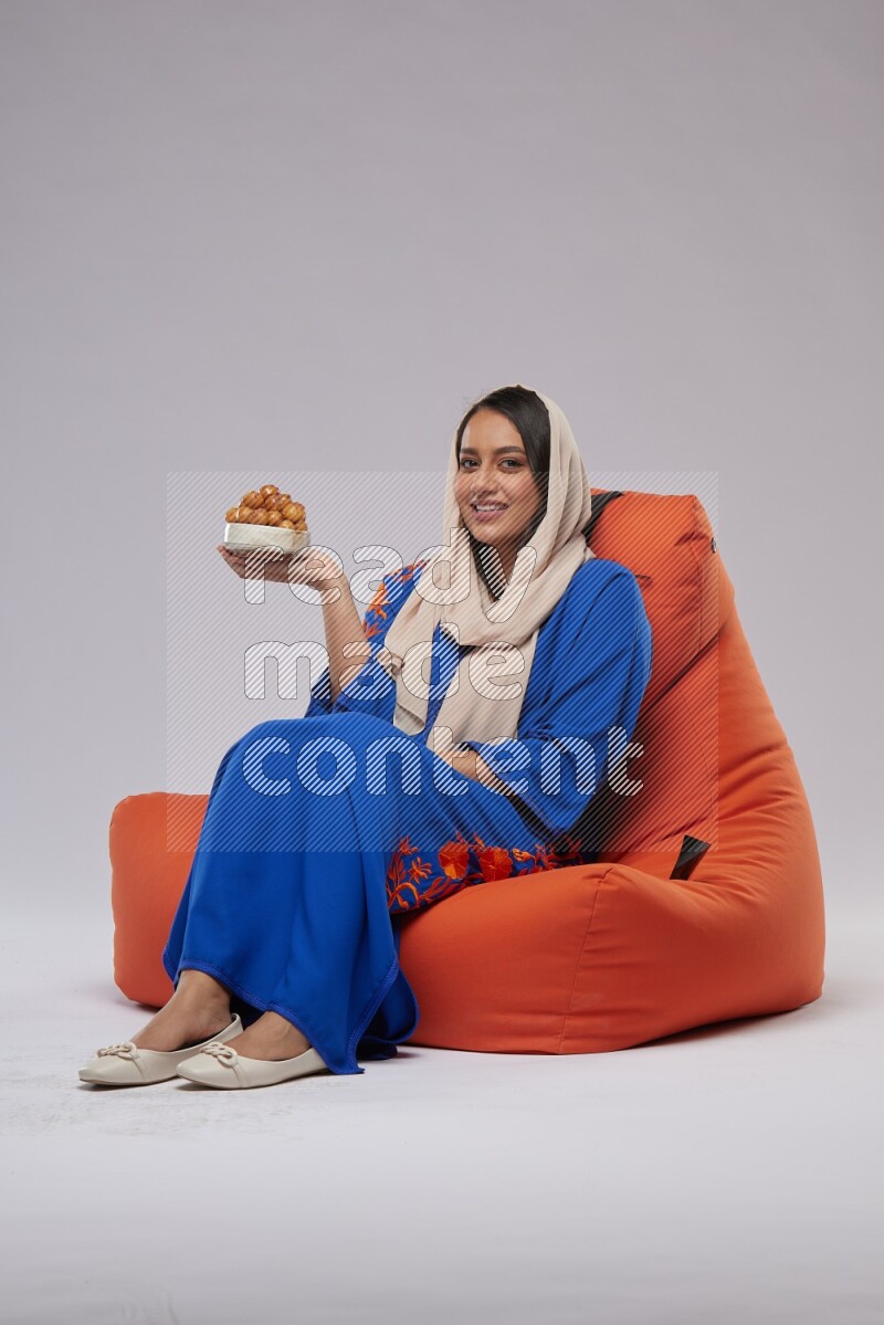 A Woman sitting on an orange beanbag wearing Jalabeya holding a plate of luqaimat