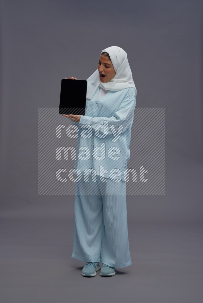 Saudi woman wearing hijab clothes standing showing tablet to camera on gray background