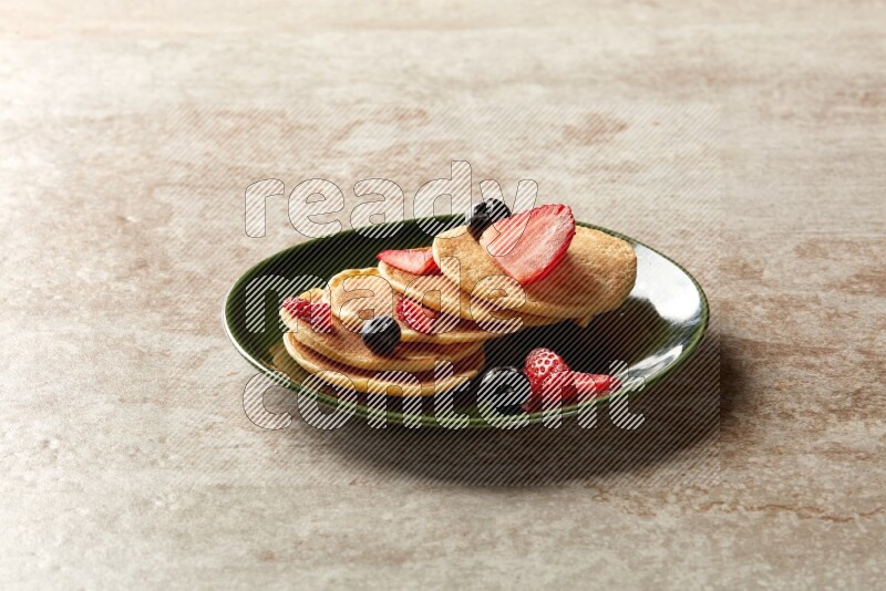 Five stacked mixed berries mini pancakes in a green plate on beige background