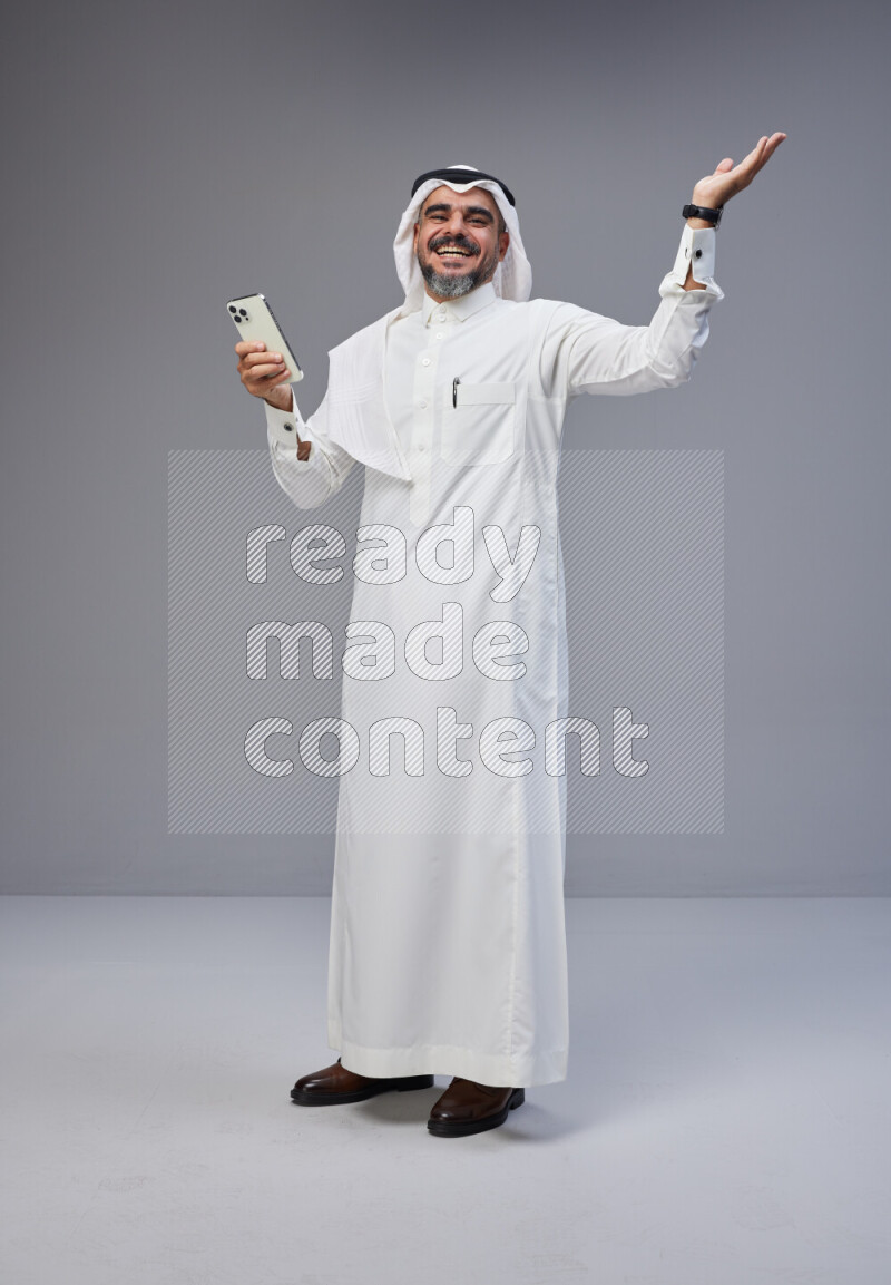 Saudi man Wearing Thob and white Shomag standing texting on phone on Gray background