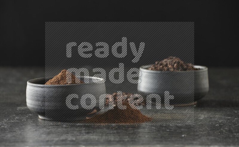2 Black pottery bowls full of cloves and the other full of cloves powder on textured black flooring