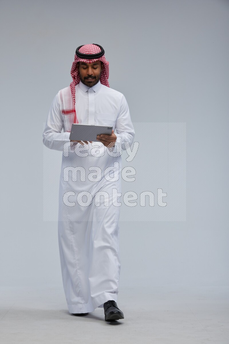 Saudi man Wearing Thob and shomag standing working on tablet on Gray background