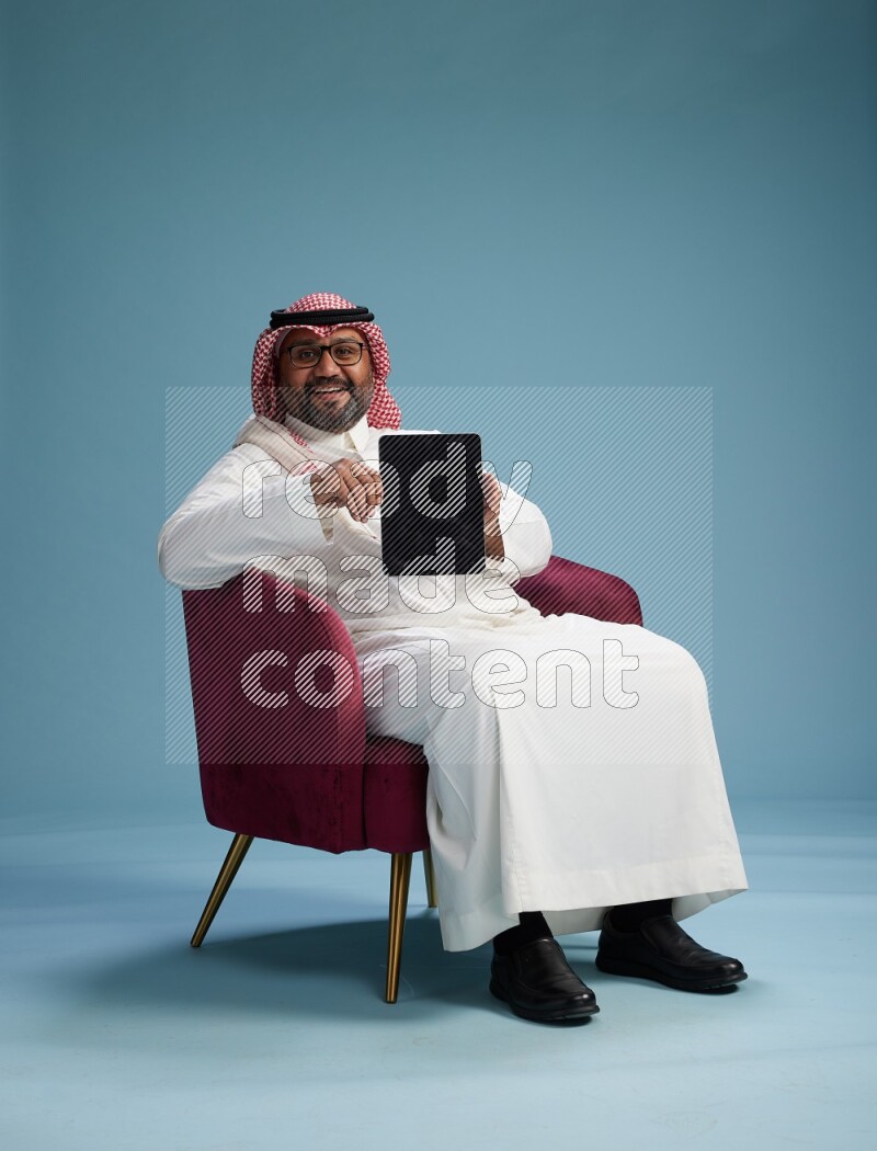 Saudi Man with shimag sitting on chair working on tablet on blue background