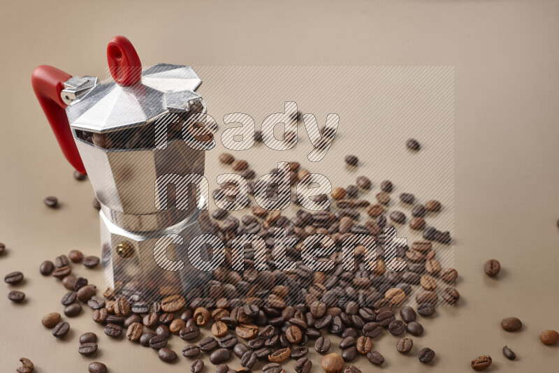A moka pot with red handle surrounded by roasted coffee beans on beige background