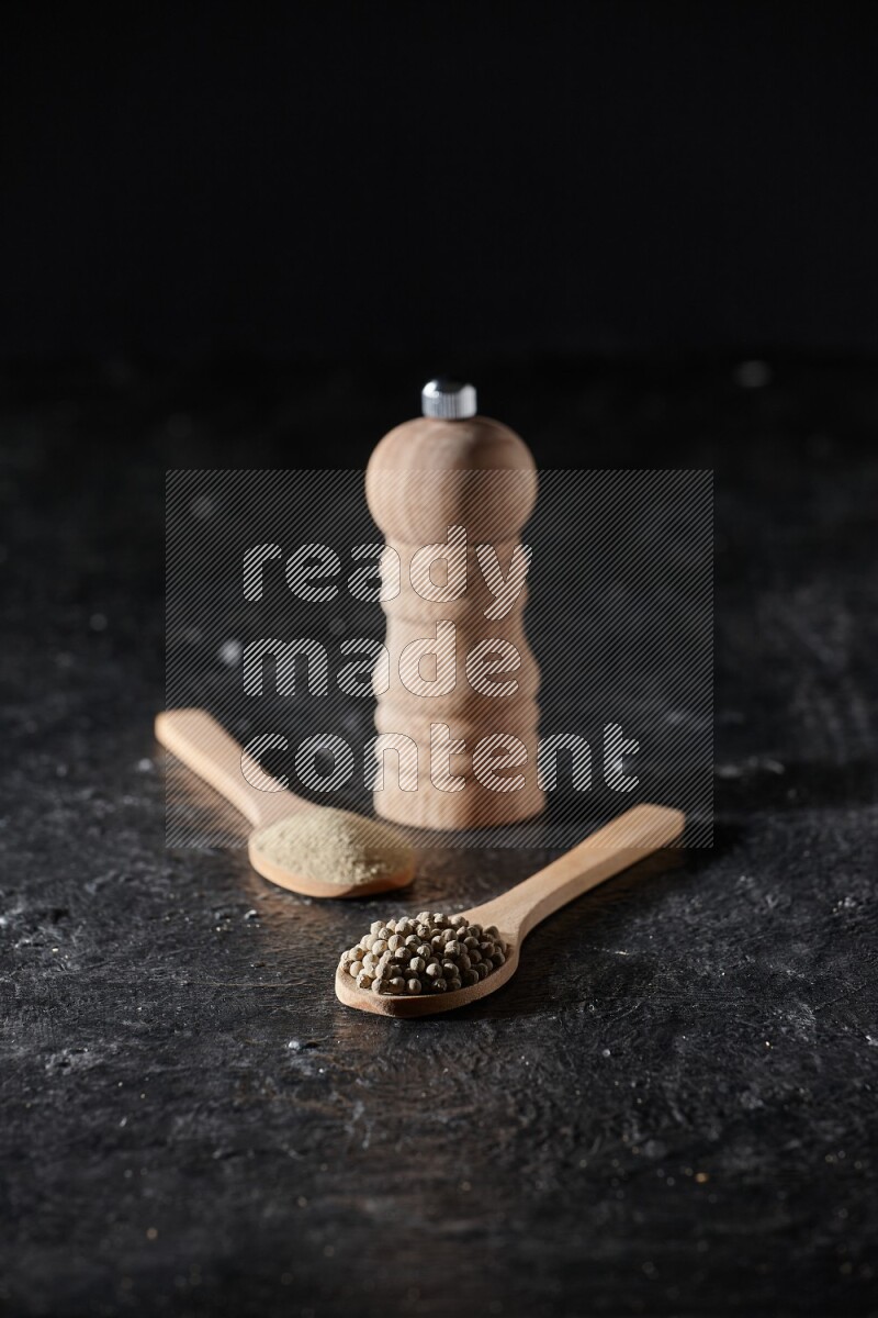 2 wooden spoons one full of white pepper powder and the other with pepper beads and a wooden pepper mill on textured black flooring