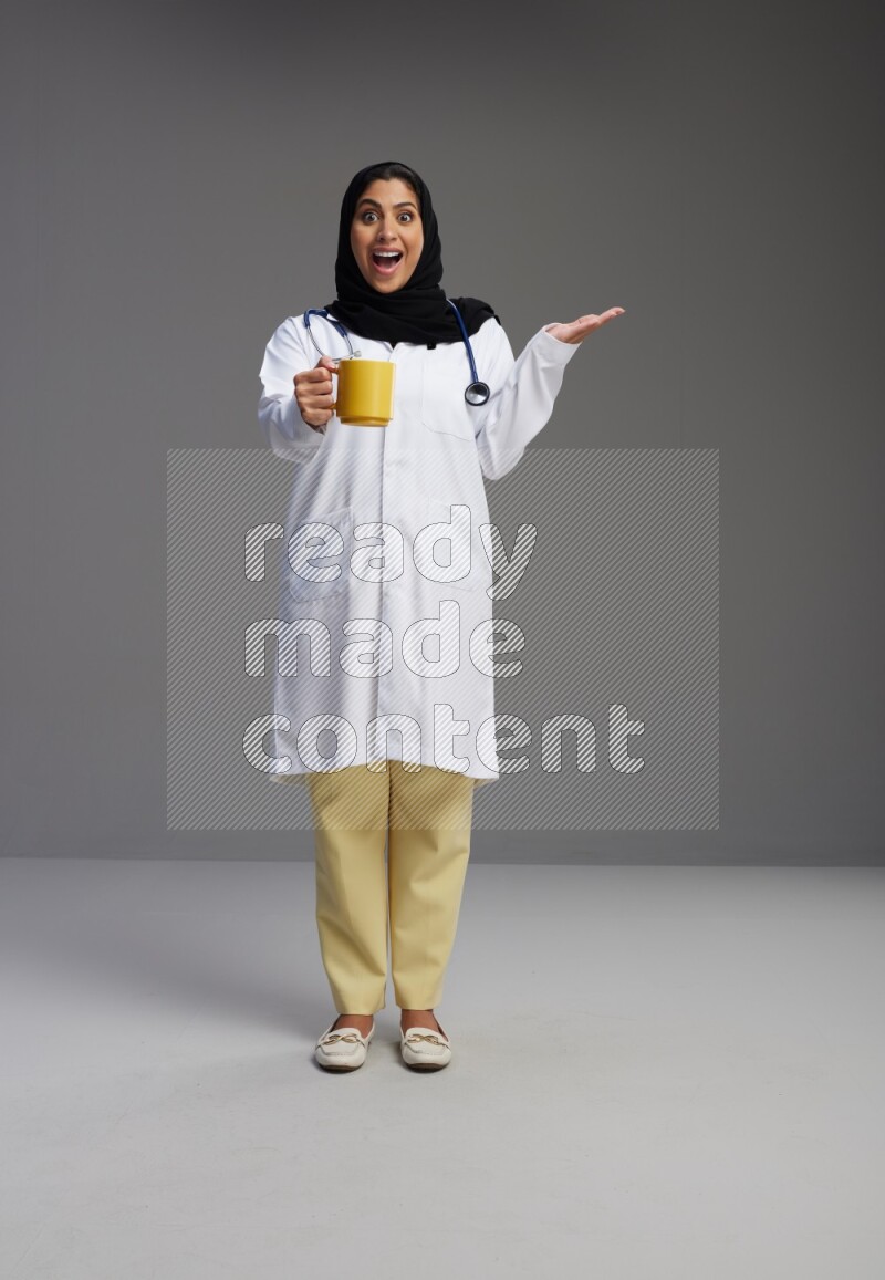 Saudi woman wearing lab coat with stethoscope standing holding mug on Gray background