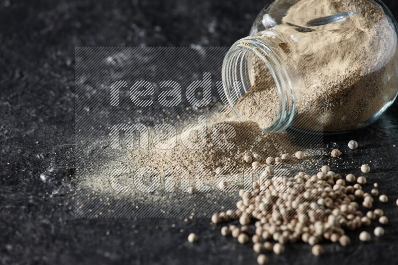 A flipped herbal glass jar full of white pepper powder with spilled powder and pepper beads on textured black flooring