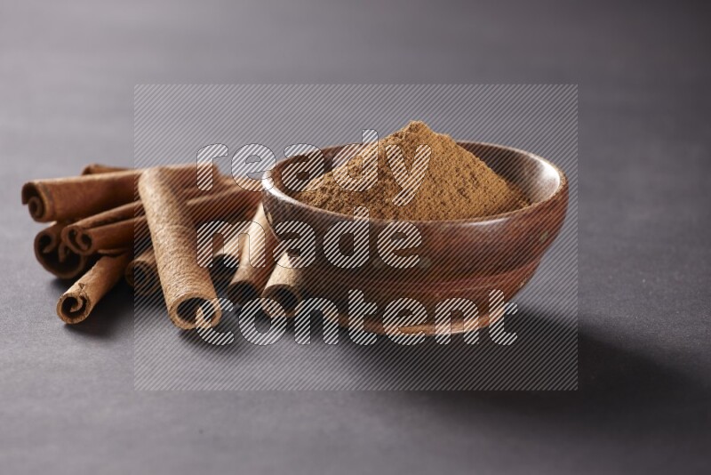 Cinnamon sticks stacked beside a wooden bowl full of cinnamon powder on black background
