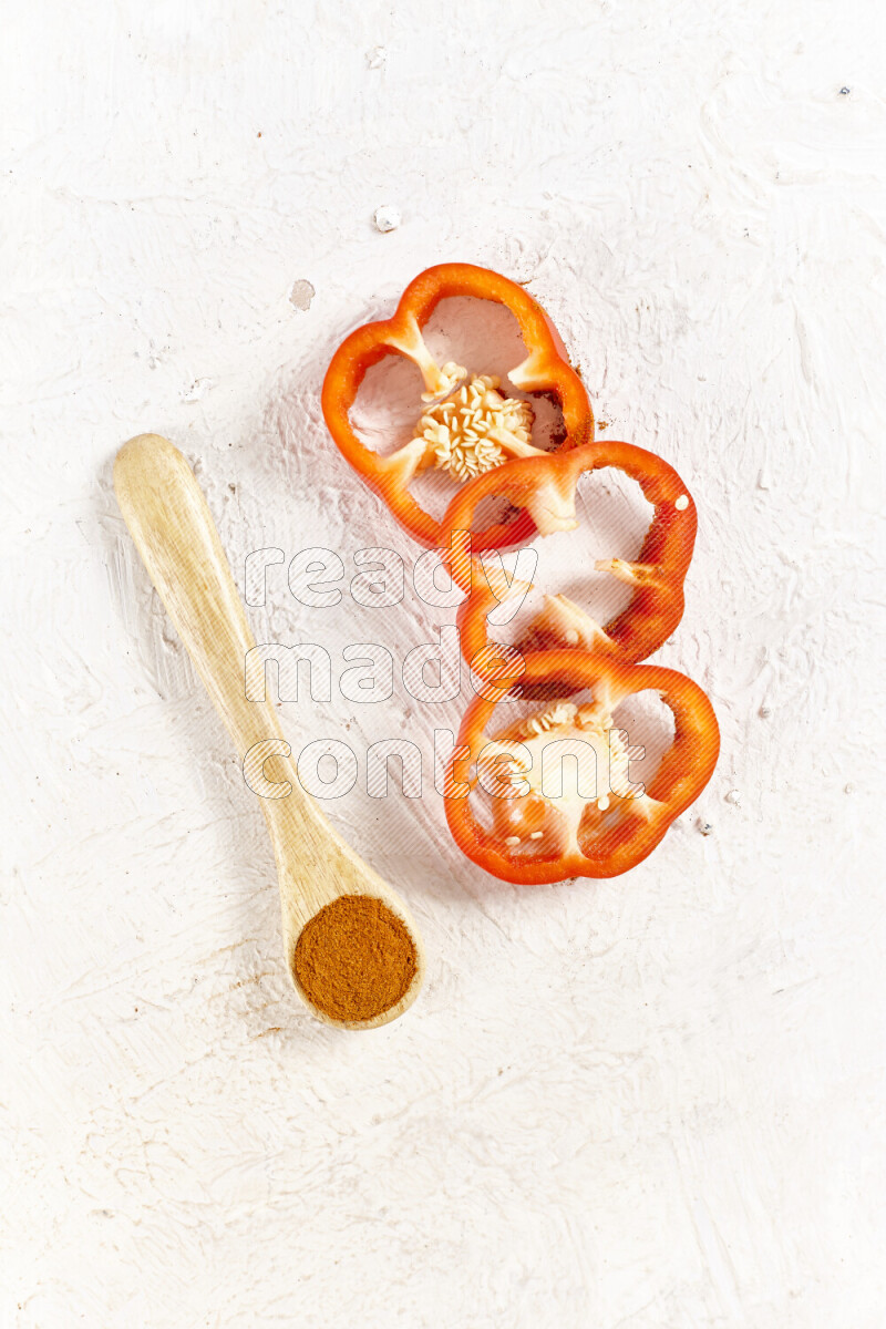 A wooden spoon full of ground paprika powder with red bell pepper slices beside it on white background