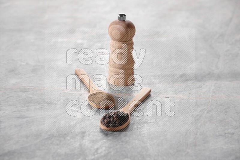2 wooden spoons, one full of black pepper powder and the other full of black pepper beads and wooden grinder on a marble flooring