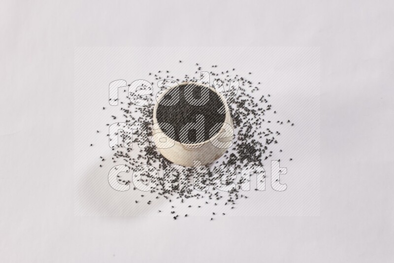 وعاء بيج فخاري ممتلئ ببذور حبة البركة مع المزيد من البذور المتناثرة علي خلفية بيضاء