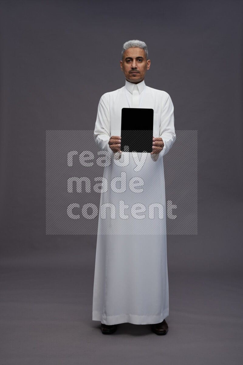 Saudi man wearing thob with neck strap employee badge standing showing tablet to camera on gray background