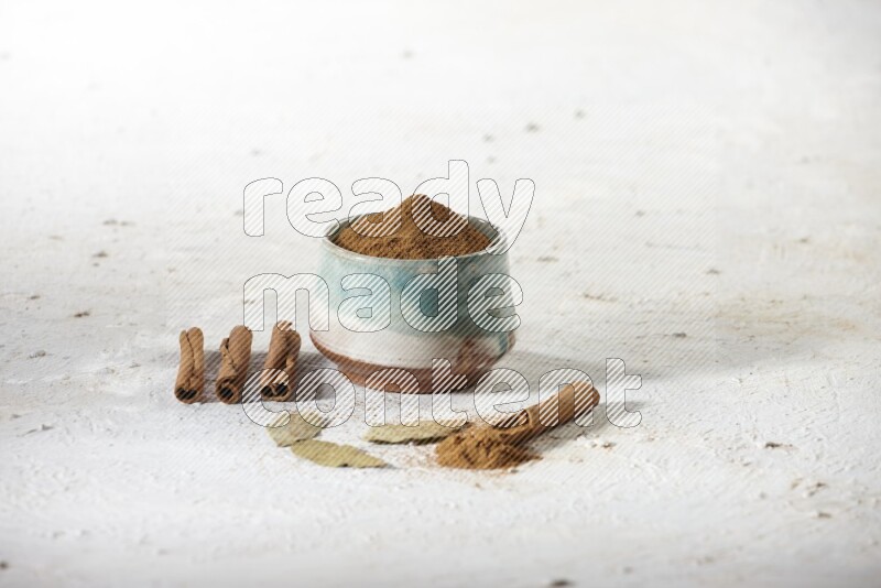Cinnamon powder in a ceramic bowl with cinnamon sticks and laurel leaves on white background