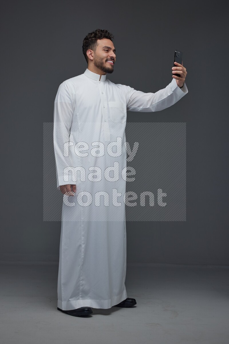 A Saudi man wearing Thobe and taking selfie with his phone making different poses eye level on a gray background