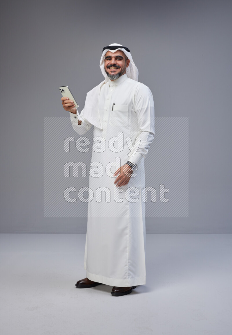 Saudi man Wearing Thob and white Shomag standing texting on phone on Gray background