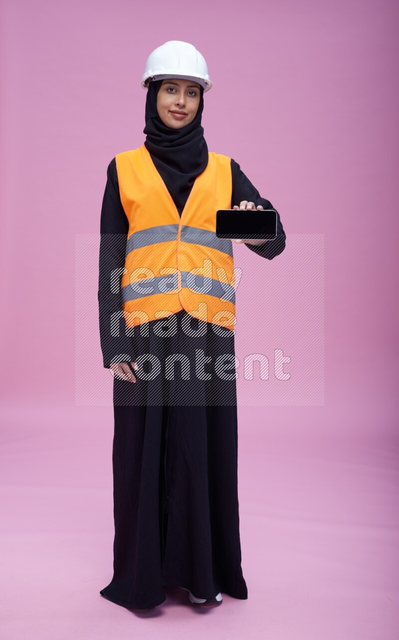Saudi woman wearing Abaya with engineer vest and helmet standing showing phone to camera on pink background