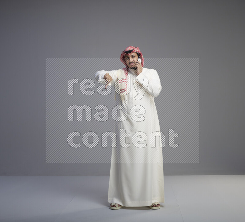 A Saudi man standing wearing thob and red shomag talking on phone on gray background