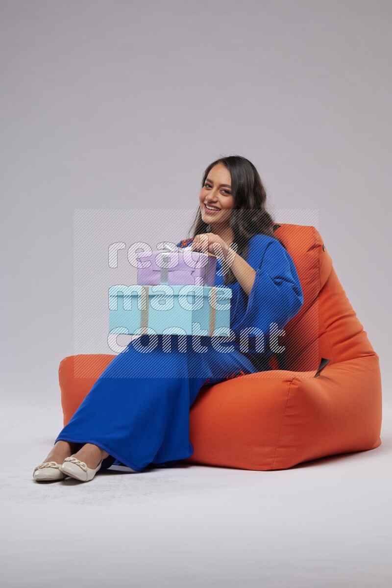 A woman sitting on an orange beanbag wearing Jalabeya holding a gift box