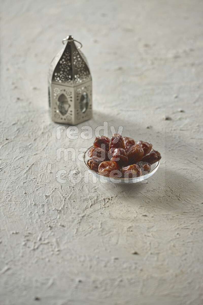 A silver lantern with different drinks, dates, nuts, prayer beads and quran on textured white background