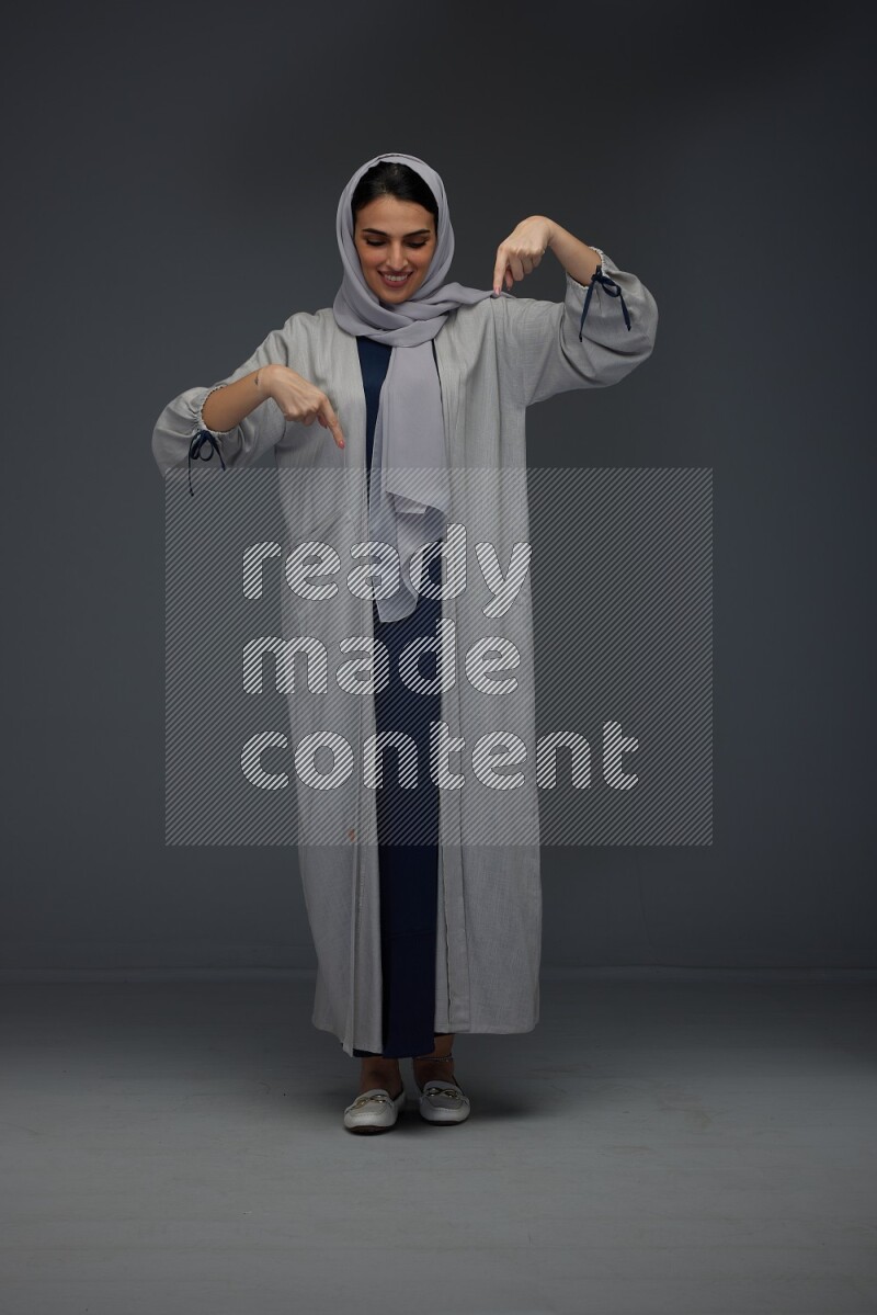 A Saudi woman wearing a light gray Abaya and head scarf standing and pointing in different directions eye level on a grey background