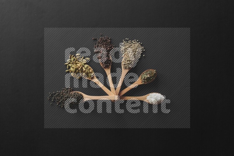 wooden spoons filled with white peppers, cloves, cardamom, salt, black peppers and basil on black flooring