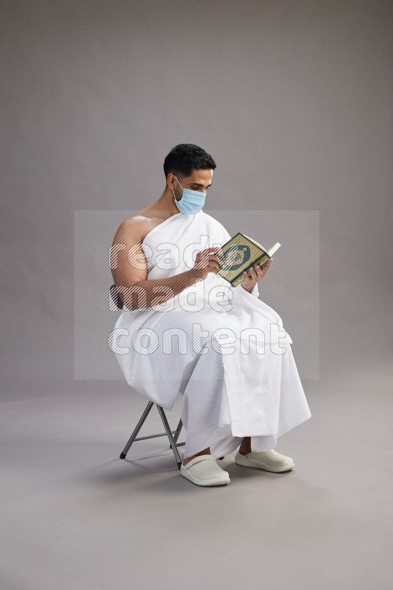 A man wearing Ehram with face mask sitting on chair reading quran on gray background
