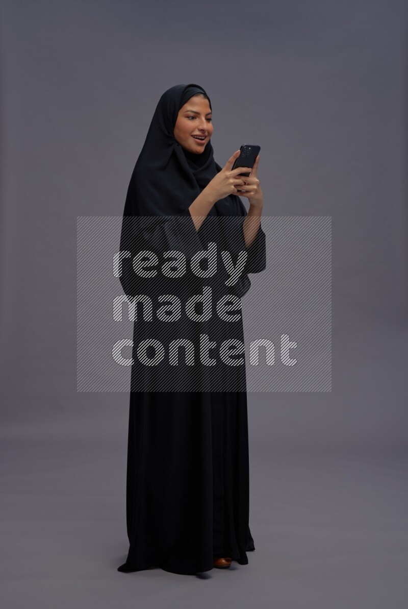 Saudi woman wearing Abaya standing texting on phone on gray background