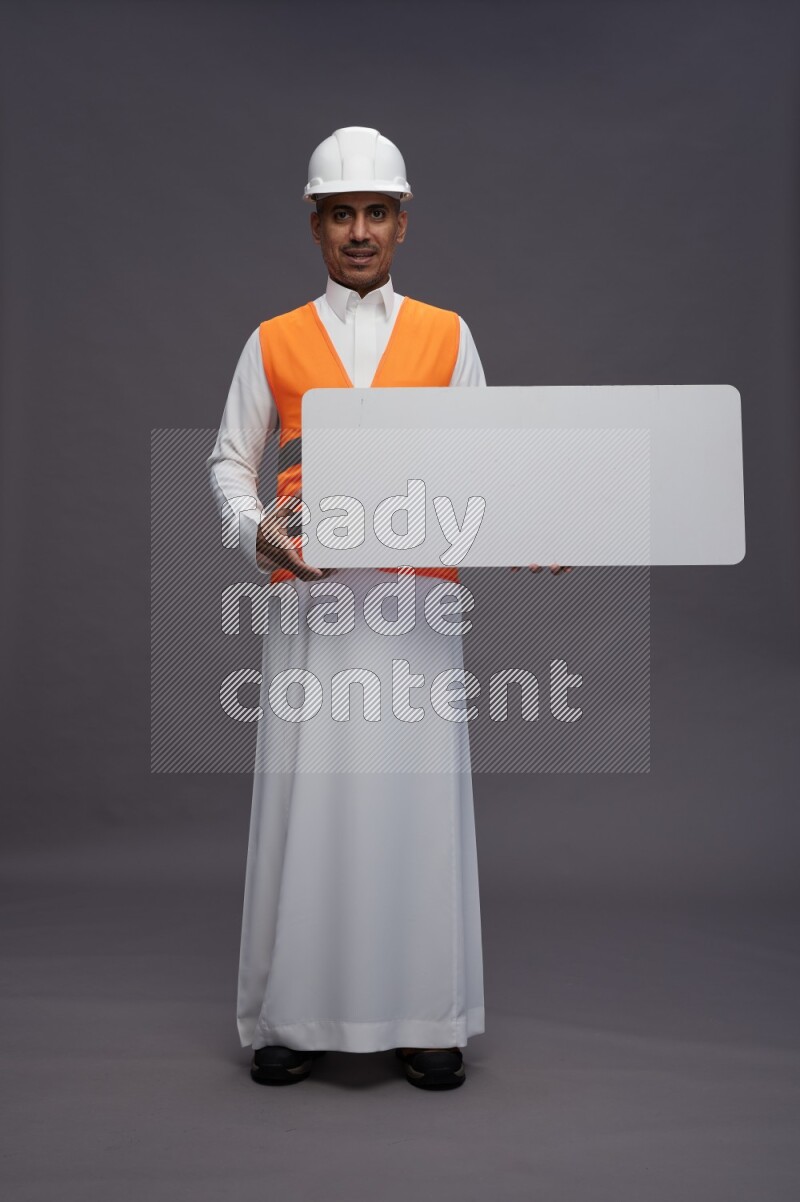 Saudi man wearing thob with engineer vest standing holding board on gray background
