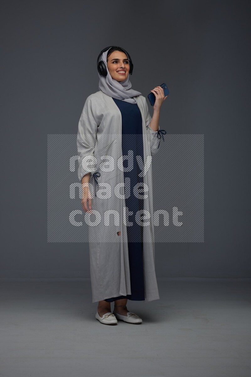 A Saudi woman wearing a light gray Abaya and head scarf standing and listening to music on a grey background