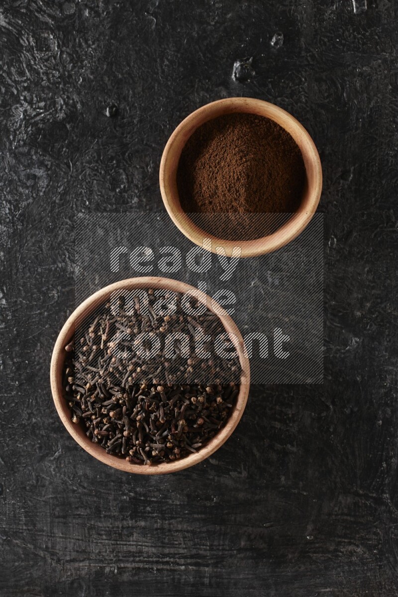2 wooden bowls full of cloves powder and whole cloves on a textured black flooring