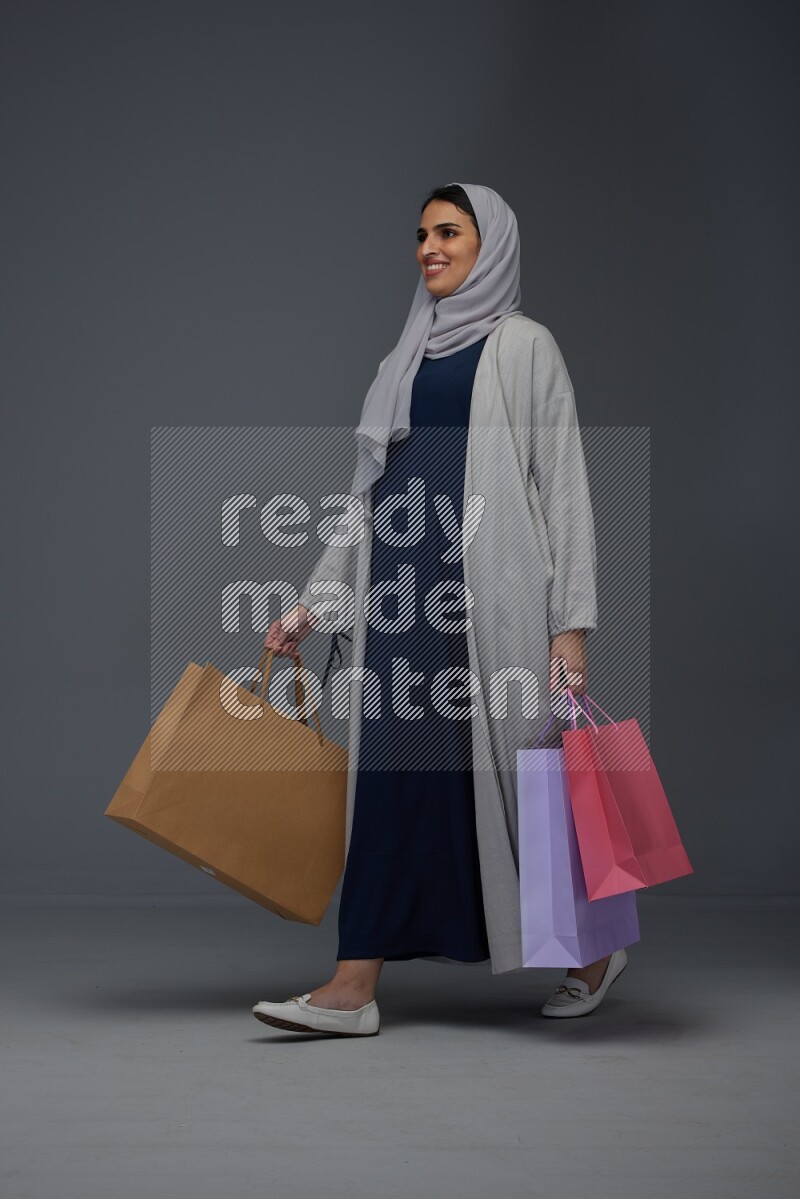 A Saudi woman wearing a light gray Abaya and head scarf standing and holding shopping bags on a grey background