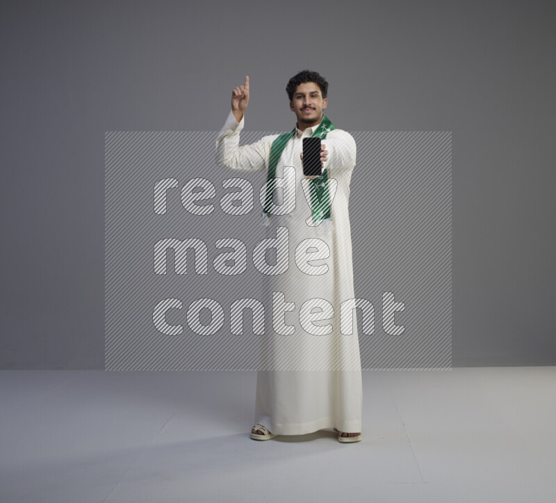 A Saudi man standing wearing thob and Saudi flag scarf showing phone to camera on gray background