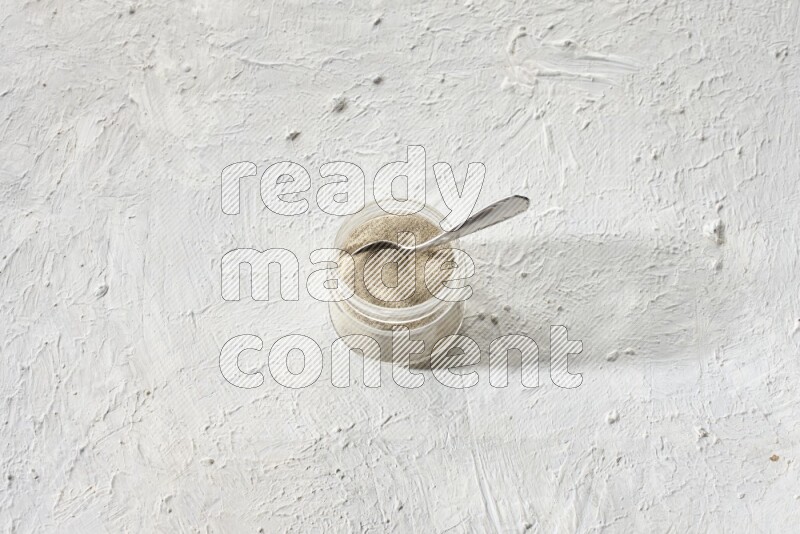 A glass jar and a metal spoon full of white pepper powder on textured white flooring