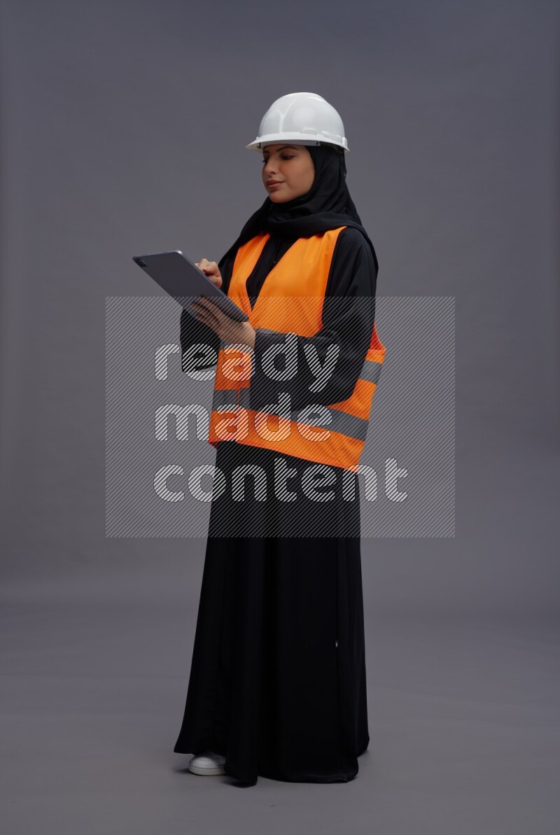 Saudi woman wearing Abaya with engineer vest standing working on tablet on gray background