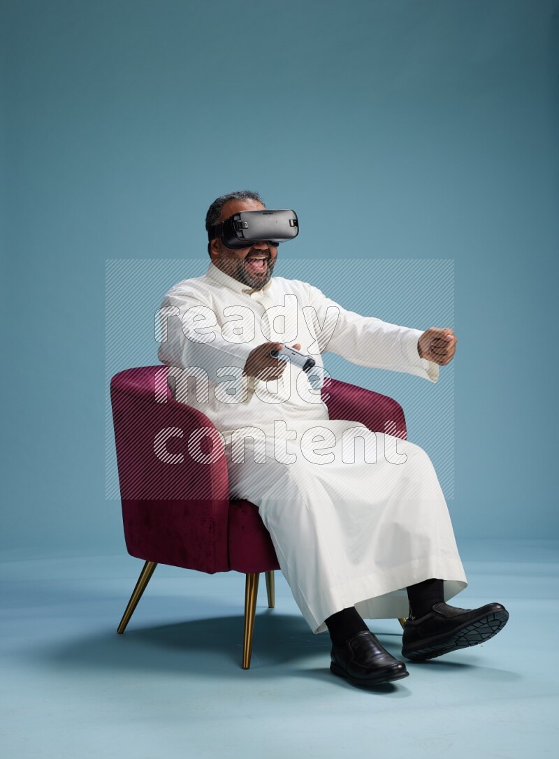 Saudi Man without shimag sitting on chair  gaming with VR and joystick on blue background