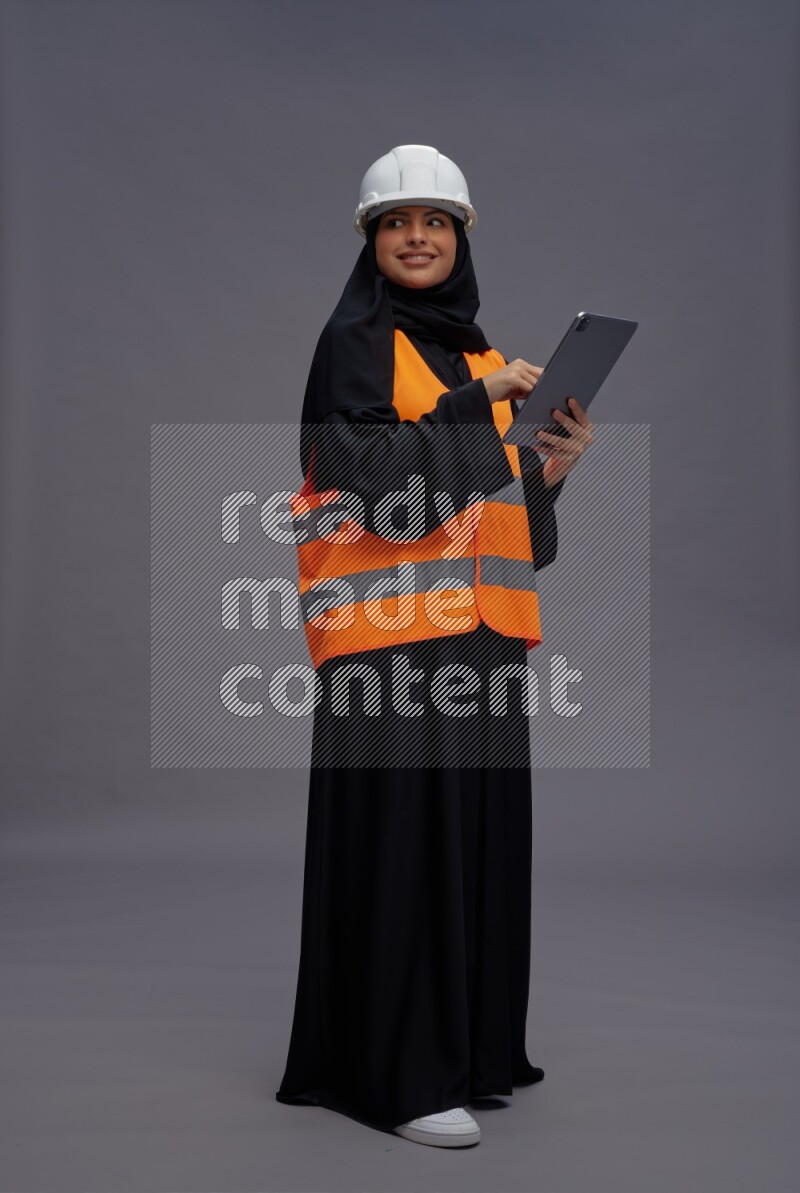 Saudi woman wearing Abaya with engineer vest standing working on tablet on gray background