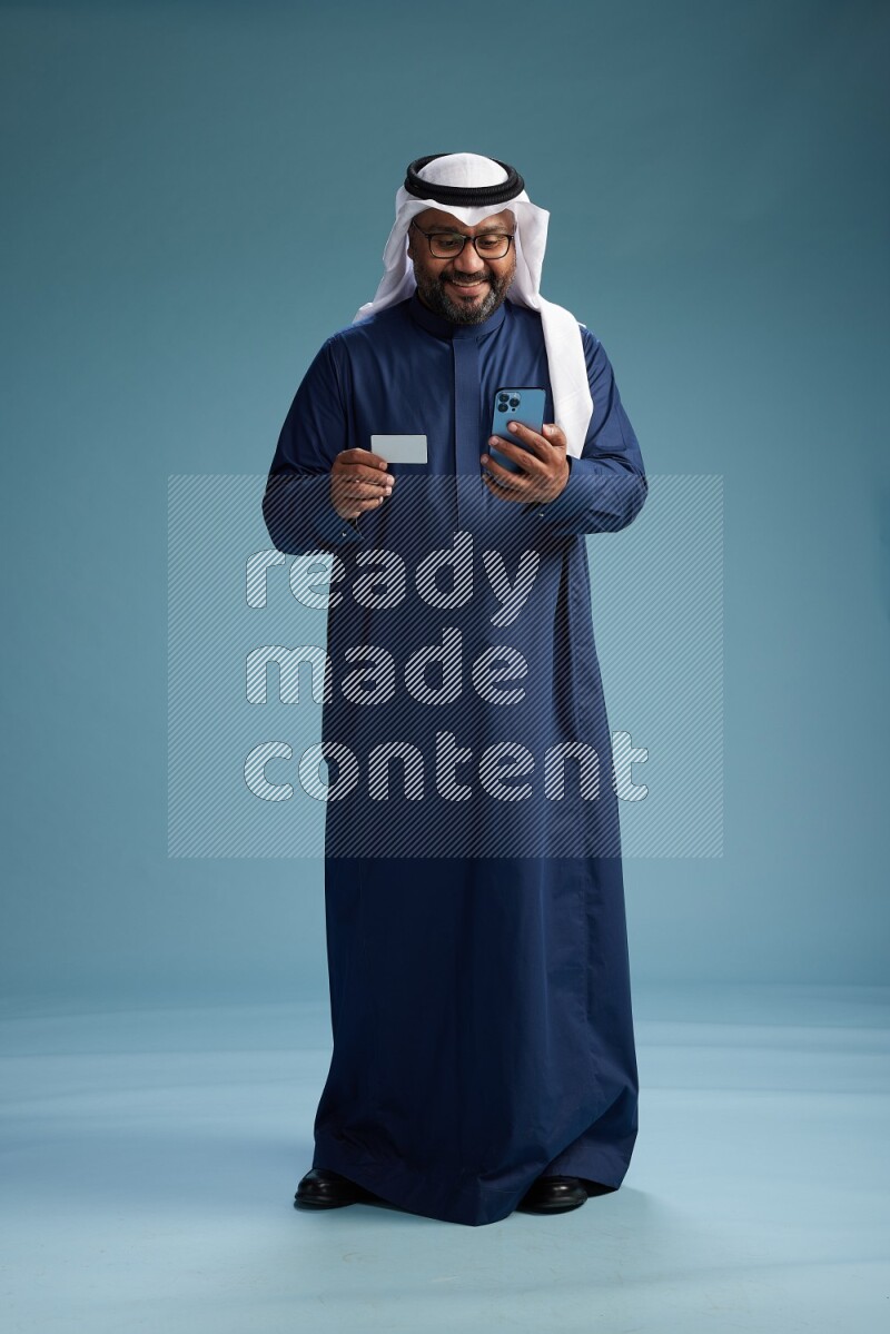 Saudi Man with shimag Standing holding ATM card while talking on phone on blue background