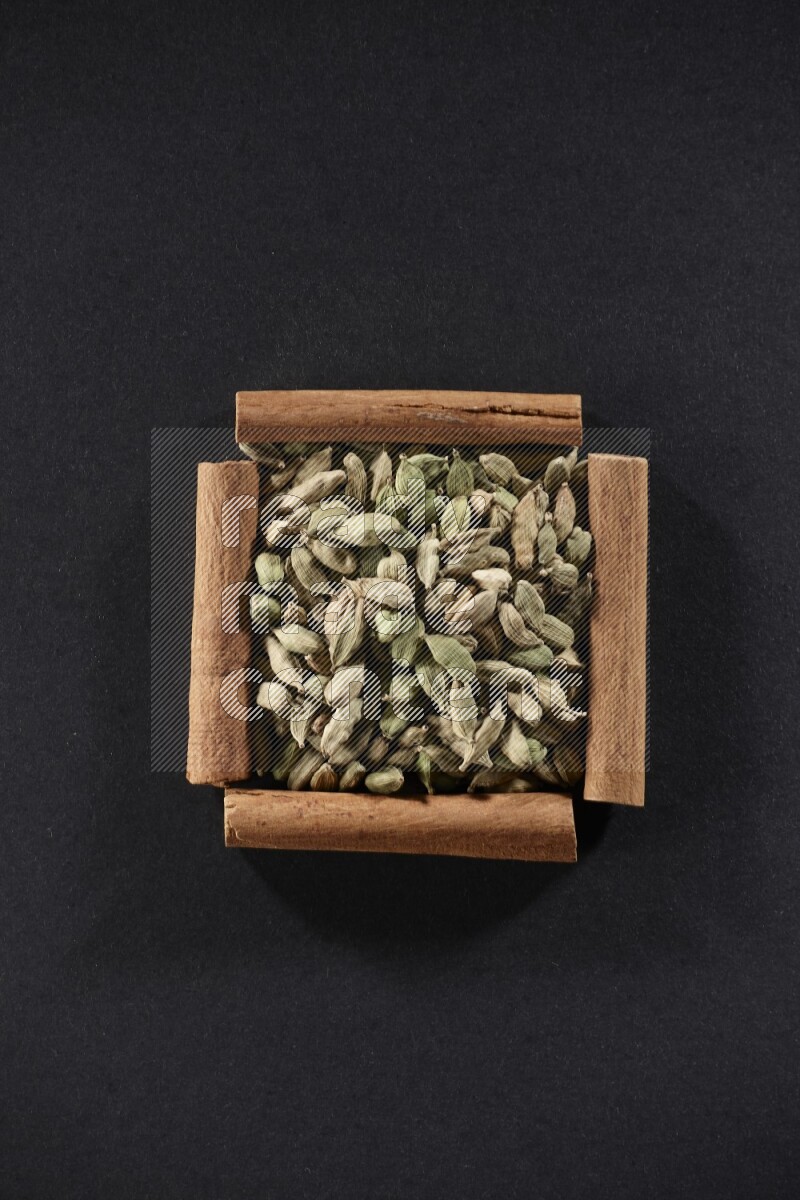 أربعة من أعواد القرفة مكونة شكل مربع بداخلها حبوب الهيل علي خلفية سوداء